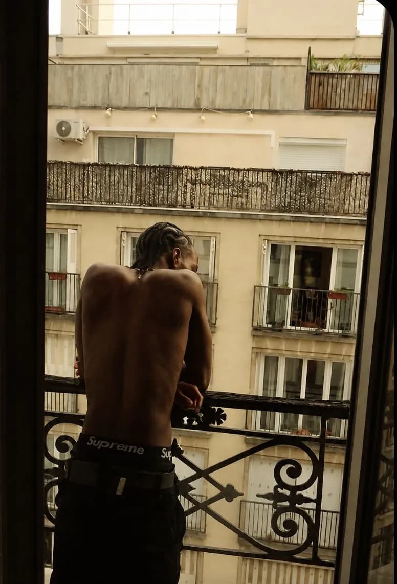 A shirtless man with braided hair stands at an open balcony door, looking out at the apartment building across the street in warm evening light.